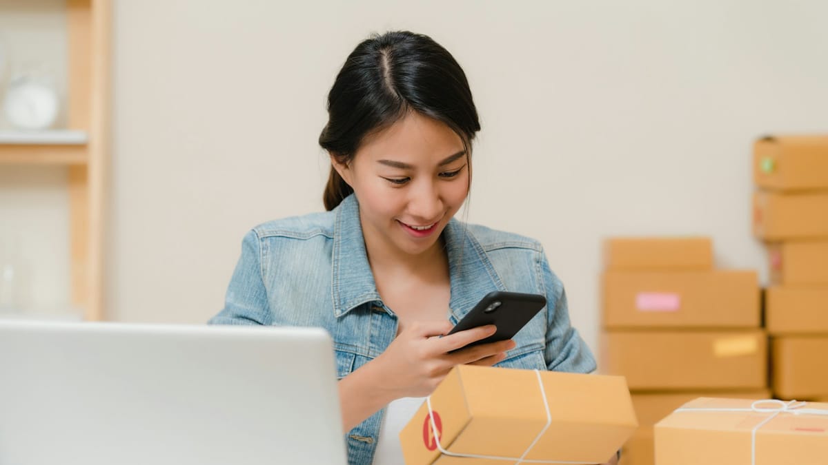 Store owner checking an order on her phone beside shipping boxes